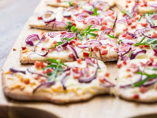 Flammkuchen-Ideen: Dünner Teig mit Käse, roten Zwiebeln, Speck und Rucola belegt. Perfekt für den Sommer!