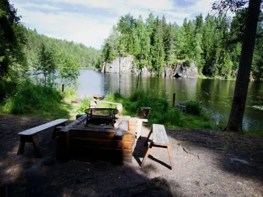 En mysig eldstad med bänkar vid en stilla sjö, omgiven av grönskande skog. Ett perfekt utflyktsmål i Dalarna för avkoppling.