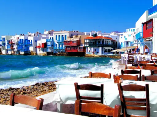 Cosa vedere a Mykonos in crociera: case colorate sul mare, onde che si infrangono sulla spiaggia di ciottoli e tavoli apparecchiati per godersi il panorama.