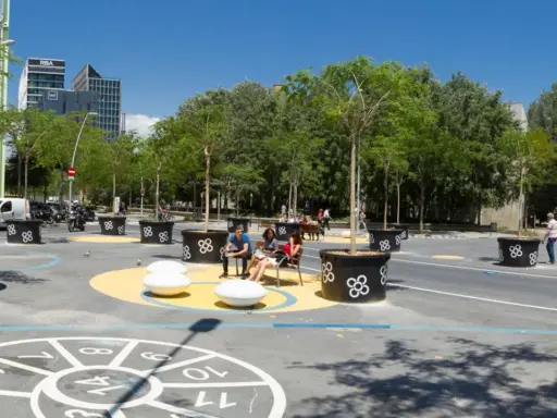 Barcelona autofrei: Menschen genießen einen grünen Platz mit Bäumen in großen Kübeln, Sitzgelegenheiten und einem Spielbereich auf dem Boden.