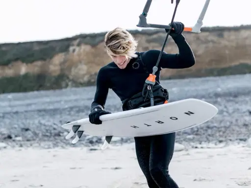 Młody kitesurfer w czarnym piance, doceniając jej właściwości termoizolacyjne, przygotowuje deskę do wodnych szaleństw.