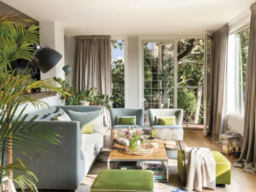 Sofá gris y otomanas verdes crean un ambiente acogedor. Plantas exuberantes y luz natural inundan el salón.