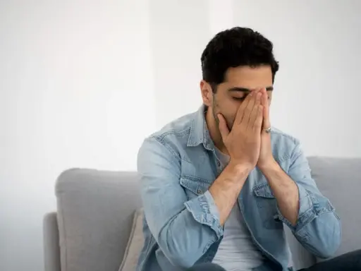 Mann sitzt auf Sofa, Hände vors Gesicht gepresst, Augen geschlossen. Fühlt sich nach dem Rauchen schwindelig.