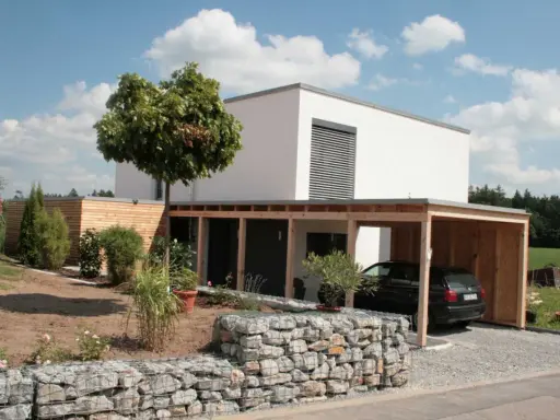 Modernes, weißes Haus mit Holzcarport und Gabionenmauer. Ein Beispiel, wie man ein low budget haus bauen kann, das dennoch stilvoll aussieht.
