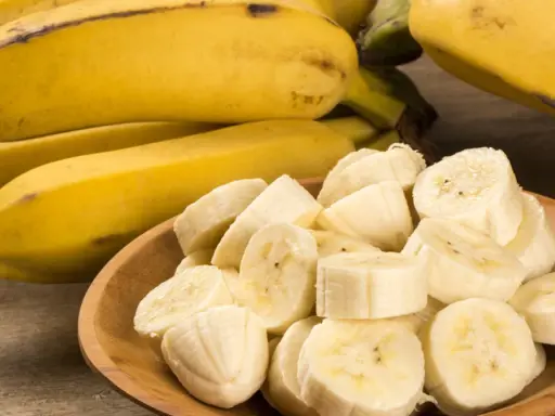 Kawałki bananów w drewnianej misce obok całych owoców. Te żółte banany, bogate w potas, są świetne dla zdrowia nerek.