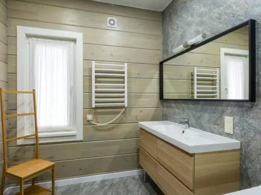 Salle de bain moderne avec murs en bois clair et pierre grise. Un meuble vasque en bois, un miroir, un radiateur et une échelle en bambou pour habiller un mur.