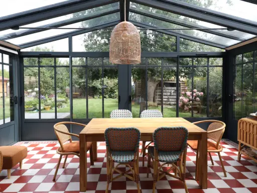 Salle à manger lumineuse dans une véranda avec toiture zinc. Table en bois, chaises colorées, sol à damier rouge et blanc, vue sur jardin verdoyant.