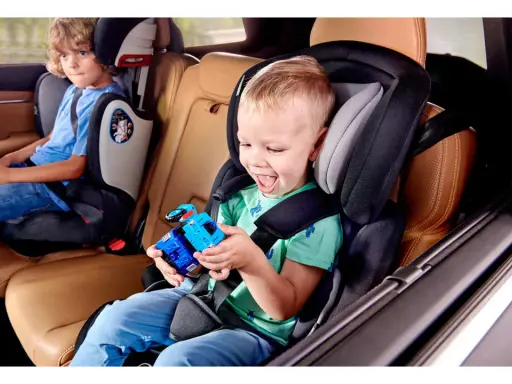 Uśmiechnięty chłopiec bawi się zabawką w foteliku Kinderkraft Comfort Up ADAC. Obok siedzi starszy brat.