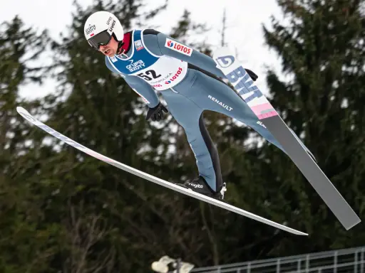 Skoczek narciarski w locie, numer 52, podczas kwalifikacji w skokach. W tle drzewa.