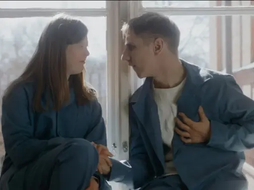 Två unga människor, klädda i blåa arbetskläder, sitter vid ett fönster. Scen från filmen om vipeholmsexperimenten.