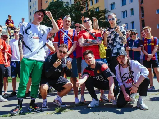 Grupa młodych ludzi w strojach piłkarskich, pozująca do zdjęcia. W tle budynki mieszkalne. To świetny **reto tekst** dla fanów futbolu.