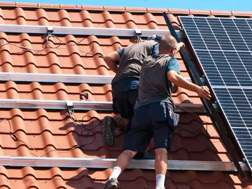 Två personer arbetar med att montera solceller på ett tegeltak. De fäster solpanelerna på monteringsskenor.