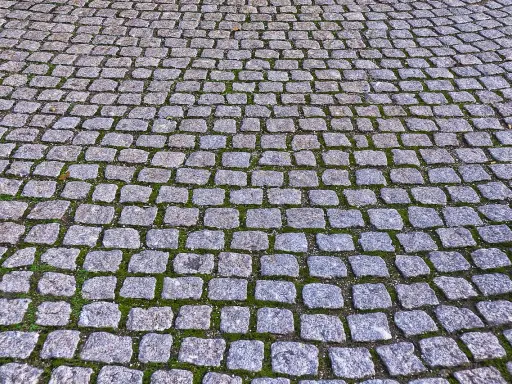 Szara kostka granitowa ułożona na ścieżce, z widocznym zielonym mchem między kamieniami. Fugowanie kostki granitowej suchym betonem zapewnia trwałość nawierzchni.
