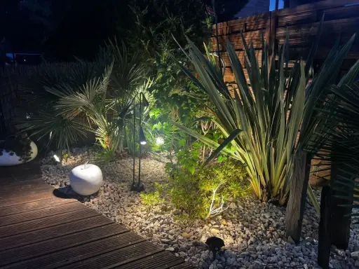 Jardin illuminé la nuit, créant une ambiance chaleureuse. La **norme éclairage extérieur** est respectée pour mettre en valeur la végétation luxuriante et le chemin en bois.