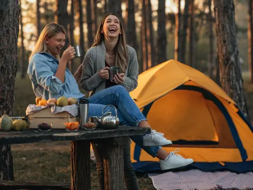 Dwie kobiety śmieją się na kempingu, siedząc przy stole z owocami i napojami. Obok rozstawiony jest żółty namiot.