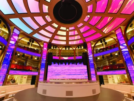 Interni di una nave da crociera, con un palco illuminato da luci rosa e blu, che ricorda la costa mediterranea.