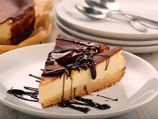 Kawałek prostego sernika z serka mascarpone, polany czekoladą, na białym talerzu.