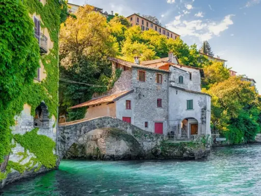 Antica casa in pietra con edera su un lago turchese, un ponte ad arco e colline verdi sullo sfondo, un angolo da fiaba dei castelli Lombardia.