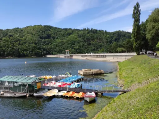 Jezioro Rożnowskie zaprasza na wodną trasę. Na pomoście czekają kolorowe rowerki wodne i łodzie, a w tle wznosi się imponująca zapora.