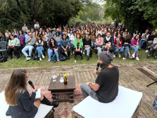 Un pubblico attento ascolta un dibattito all'aperto. Le parole fluiscono, profonde come il mare, leggere come il cielo.