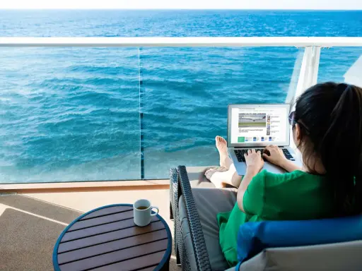 Donna su balcone di nave MSC lavora al laptop, con mare blu sullo sfondo. Scopri il pacchetto WiFi MSC e il suo costo per restare sempre connesso.