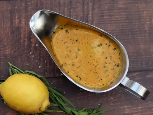 Eine cremige Cafe de Paris Sauce in einer Sauciere, daneben ein Zitrone und Kräuter auf dunklem Holz.