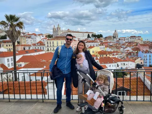 Familia disfrutando de las vistas de Lisboa con niños. Un bebé en portabebés y una niña en carrito, con la ciudad de tejados naranjas de fondo.