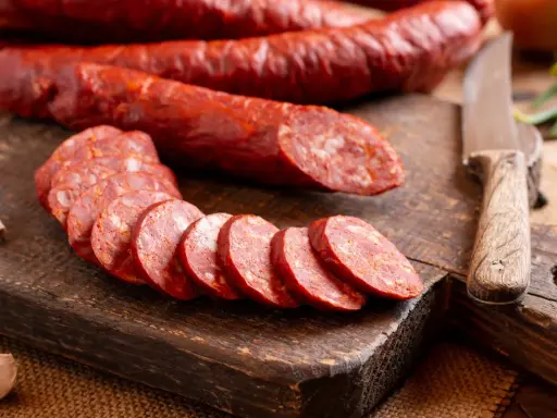 Salami auf einem Holzbrett, geschnitten und im Ganzen. Man kann Salami einfrieren, um sie länger haltbar zu machen. Knoblauch und Messer liegen daneben.