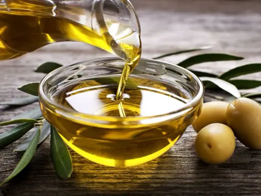 Aceite de oliva virgen extra vertiéndose en un cuenco de cristal, rodeado de aceitunas y hojas de olivo sobre madera rústica.