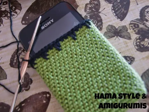Funda de móvil tejida a crochet, un patrón verde con borde negro. Un gancho y hilo negro están listos para continuar el proyecto.