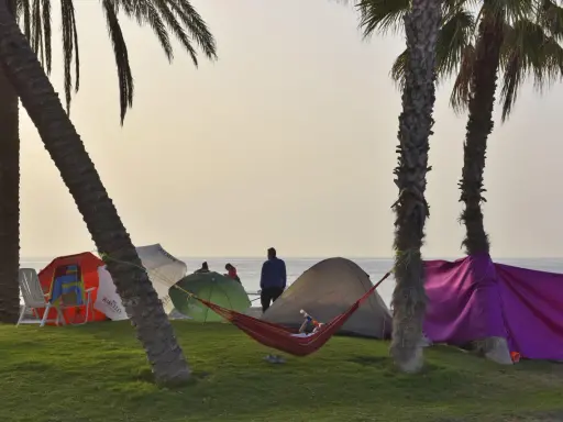Entspannter Camping-Urlaub an der Costa del Sol. Palmen, Zelte, Sonnenschirme und eine Hängematte am Strand.
