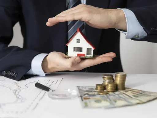 Manos protegen una casa modelo sobre un contrato de alquiler con avalista, junto a dinero y gráficos.
