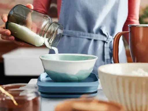 Eine Hand schüttet Mehl aus einem Glas in eine Schüssel. Die Frage, wie viel Gramm Mehl in eine Tasse passen, wird hier visuell thematisiert.