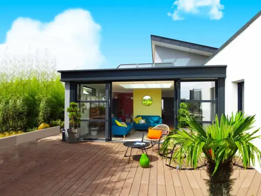 Une véranda moderne avec de grandes baies vitrées s'ouvrant sur une terrasse en bois. Un salon coloré est visible à l'intérieur.