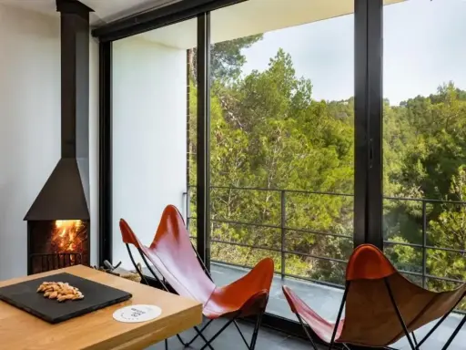 Chimenea encendida y sillones de cuero frente a un balcón con vistas a un bosque frondoso, ideal para el turismo interior España.