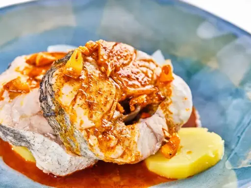 Merluza a la gallega con patatas y salsa de pimentón, servida en un plato azul.