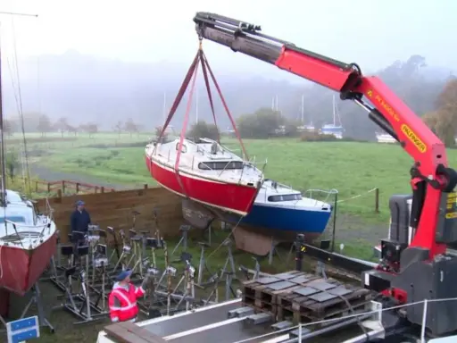 Grue soulevant un bateau rouge pour son recyclage. Un autre bateau bleu est posé sur des supports. L'opération de recyclage bateau se déroule dans un paysage brumeux.