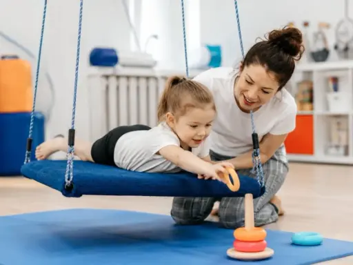 Terapia integracji sensorycznej: dziewczynka na huśtawce z terapeutką, która pomaga jej sięgnąć po zabawkę.