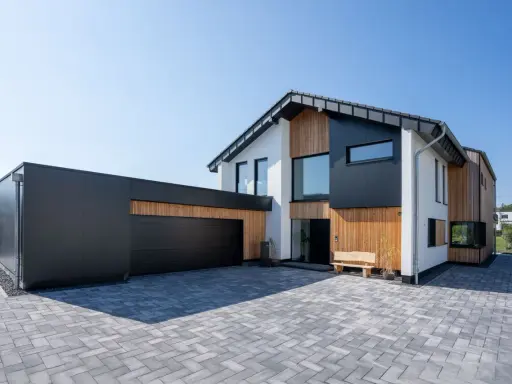 Modernes Haus mit Garage, dessen Satteldach modern interpretiert ist. Holz und dunkle Fassadenelemente prägen das Bild.