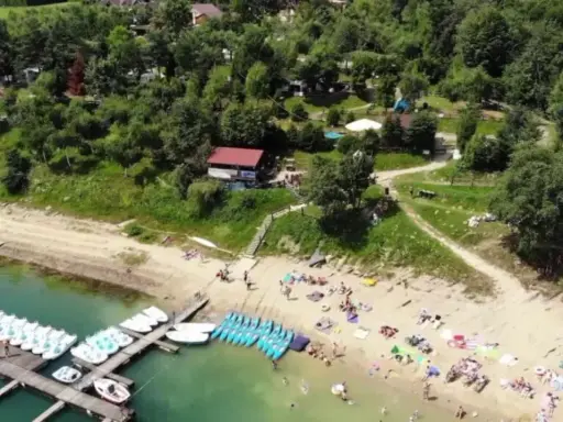 Słoneczna plaża w Polańczyku tętni życiem. Ludzie odpoczywają na piasku, a w wodzie pływają rowery wodne i kajaki.