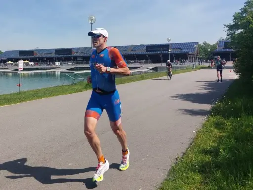Triathlet im blauen Anzug läuft auf einer Straße. Er fragt sich, wie wird man schneller. Ein Radfahrer ist im Hintergrund zu sehen.
