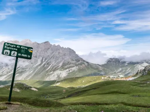 Wanderwegschild weist den Weg zum Refugio de Aliva und Espinama in den **Lagos de Covadonga**. Grüne Hügel und zerklüftete Berge unter blauem Himmel.