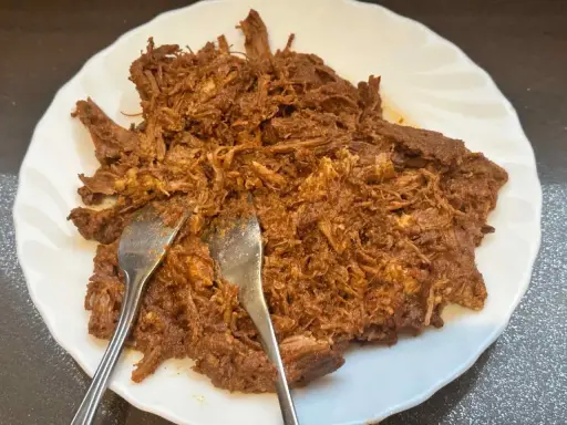Delicioso pulled pork deshebrado en un plato blanco, listo para ser servido. Dos tenedores se clavan en la carne jugosa.