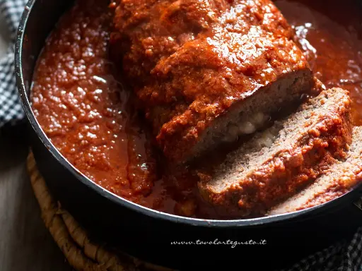 Un succulento polpettone ripieno al forno, affogato in un sugo rosso e invitante, con una fetta tagliata che rivela un cuore filante.