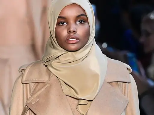 Halima Aden walks the runway in a stylish beige coat and hijab, exuding confidence and grace.