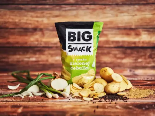 Reklamowa fotografia paczki chipsów "Big Snack" o smaku zielonej cebulki, z ziemniakami i cebulką w tle.