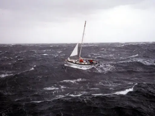 Un voilier affronte une mer agitée, sa voile blanche tendue. La mise à la cape est une manœuvre de survie face aux éléments déchaînés.