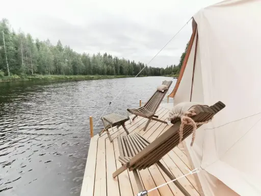 Två träfåtöljer och ett litet bord på en brygga vid en sjö, med skog i bakgrunden. En del av ett glampingtält syns. Upplev glamping Norrbotten.