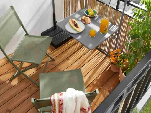 Idéal pour aménager un petit balcon en longueur : deux chaises pliantes vertes, une table avec fruits et jus, et des plantes.