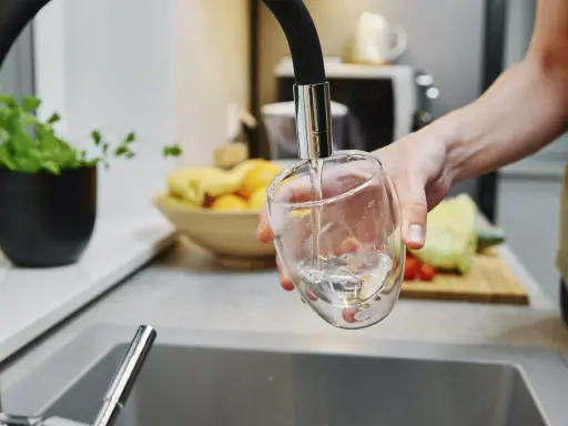 Ręka nalewa wodę do szklanki. W tle widać owoce i warzywa, jakby przygotowywano posiłek. Czysta woda, jak w akwarium.